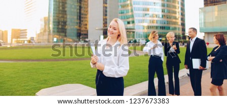 Close up face blonde secretary looking at camera and smiling with blue case in   and talking partners background. Concept of office assistant with documents outside and business employees. Beautif