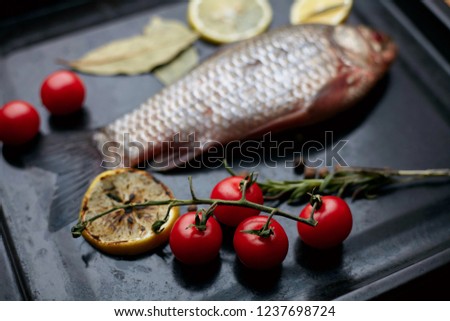 Still life of fresh silverfish on a black tray that lies on pieces of ice and around fresh cherry tomatoes, a sprig of rosemary and a fresh slice of lemon and a little toasted lemon.
