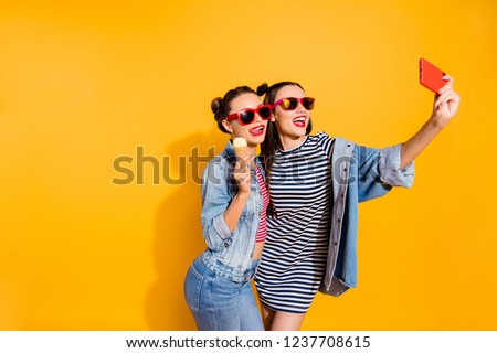 Two beautiful attractive brunette hair lady isolated on yellow background in street style stylish casual denim jeans trendy glasses spectacles take picture on cellular show tongue stick out