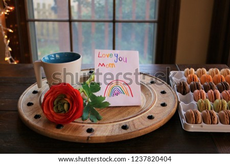 A round, wooden, rustic breakfast tray with a coffee mug, red flower, mothers day hand made card and french macrons, placed on a brown wooden table, with window in the background. 