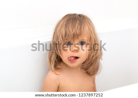 Little girl with blond wet hair laughs and shows tongue on the white backgrond. Fun bathing children. Positive hygiene. Independent child. Toddler looking at camera. Forelock covers one eye.