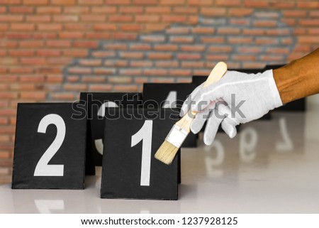 Close-up image of a white glove holding a brush near the label of a number placed on a table with a brick wall in the background.