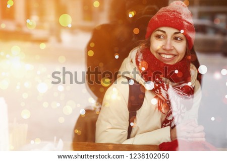 smiling happy woman in warm clothes, red knitted cap, scarf and mittens sitting in cafe near window and looking outside to the snowy street. view from outside, the window reflects snowy street