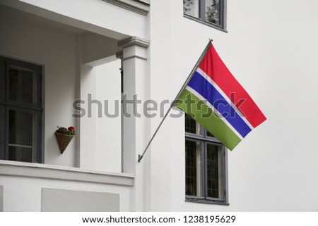 Gambia flag hanging on a pole in front of the house. National flag waving on a home displaying on a pole on a front door of a building and raised at a full staff.