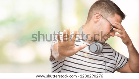 Handsome young man wearing headphones covering eyes with hands and doing stop gesture with sad and fear expression. Embarrassed and negative concept.