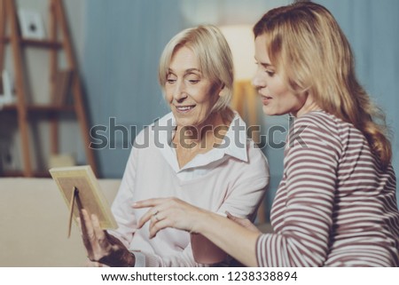 Precious memories. Adorable senior mother with young sweetheart daughter relaxing on comfortable couch and holding old photo while recalling happy moments and smiling dreamily