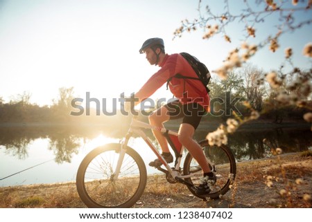 Determined young bearded man in long sleeve jersey riding a mountain bike by the lake or river. Sunset over water in background. Side view