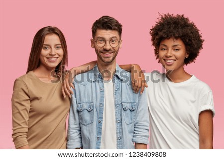 Horizontal shot of three mixed race teenagers spend time together, pose for common photo against pink background. Satisfied guy in eyewear and denim shirt stands between two cheerful women indoor Royalty-Free Stock Photo #1238409808