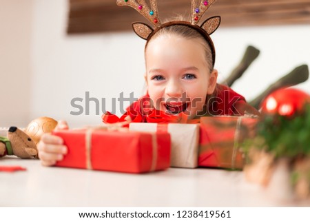 Cute young girl wearing costume reindeer antlers lying on the floor, surrounded by many christmas presents, screaming with joy. Happy kid at christmas with xmas gifts.