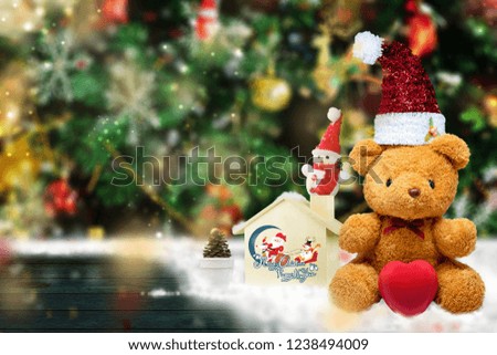 Teddy Bear wearing red hat is placed on a wooden table, with a wooden house, a snowman and a backdrop of green trees adorned with decorative items. Celebrate Christmas And happy new year