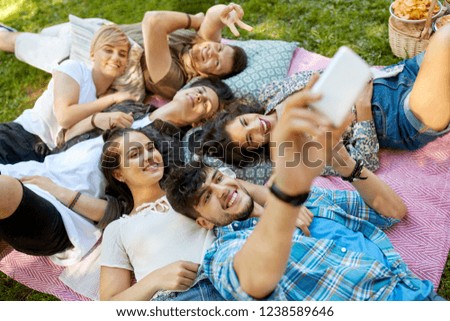 friendship, leisure, technology and people concept - group of friends chilling on picnic blanket at summer park and taking selfie by smartphones
