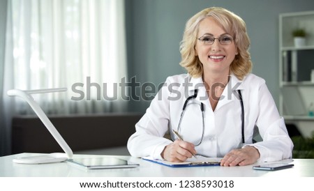 Female physician sitting at table, smiling in camera, working medical records