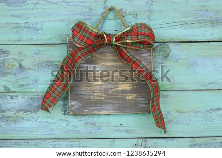 Blank wood sign with red and green plaid Christmas bow hanging on antique rustic teal blue wooden door; holiday background with aged copy space