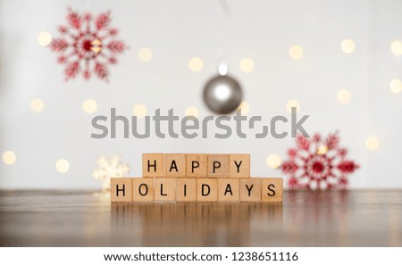 2018 Seasonal Festive Photo Banner Idea of Happy Holidays Spelled Out of Stacked Wood Block Letters with Colorful Red Snowflake Ornaments and Bright Yellow Twinkle Lights in Blurred White Background 
