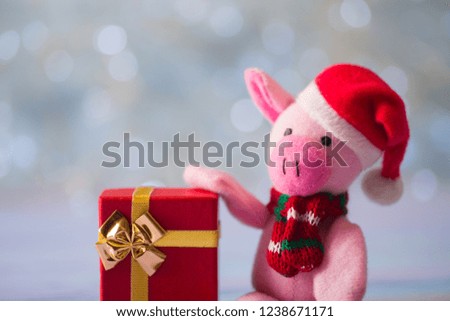 Closeup cute toy pig in Santa hat and scarf sitting on wooden table and holding festive gift box on light bokeh background. Symbol of Chinese New Year.