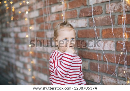 little girl blonde with blue eyes in a striped sweater on a Christmas carousel against a brick wall and luminous garland, family new year