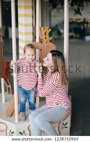 little girl blonde with blue eyes in a striped sweater and her mom on a Christmas carousel against a brick wall and luminous garland, family new year