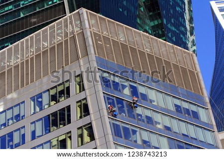 Glass cleaning workers hanging against the wall with a bucket by the support of ropes and cleaning a glass building in Moscow city. Skyscrapers background