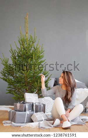 A young woman sits under a Christmas tree with gifts in a Scandinavian interior
