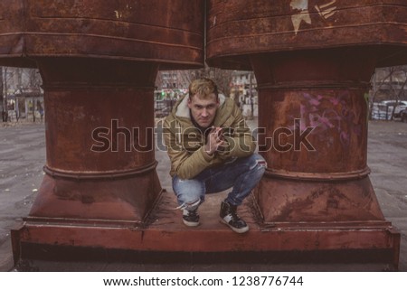 Handsome man in warm street clothes posing outside, in the background iron pipe
