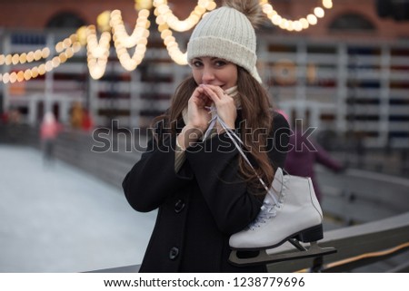 Young brunette with skates on her shoulder, on a background of winter city Park , ice rink