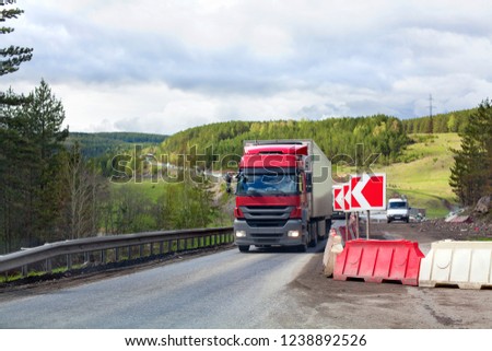 Red big truck on the road is carrying cargo, road repair, red detour sign, countryside, green forest, field, sky with clouds background