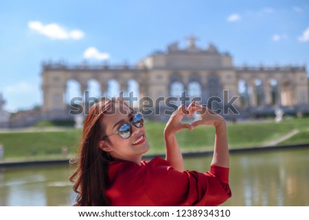 Asian tourist woman making a heart shape and smiling at camera with beautiful Schoenbrunn palace on background. Happy girl in Vienna city, Austria