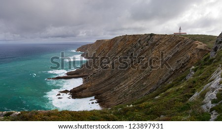 Cape Espichel cliff of sedimentary rocks, lighthouse and clouds. Sesimbra, Portugal.