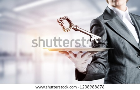 Cropped image of waitress's hand in white glove presenting big key on metal tray with office view on background.