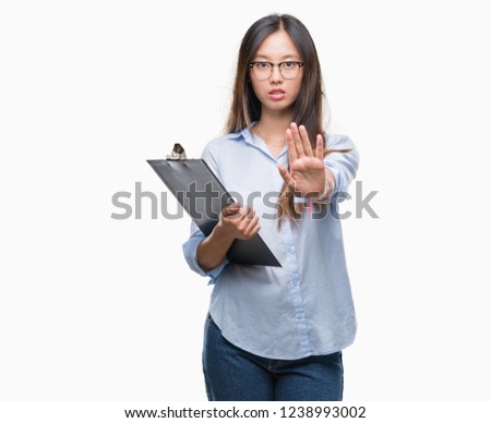 Young asian business woman holding clipboard over isolated background with open hand doing stop sign with serious and confident expression, defense gesture