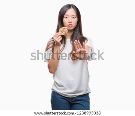 Young asian woman eating chocolate chip cookie over isolated background with open hand doing stop sign with serious and confident expression, defense gesture