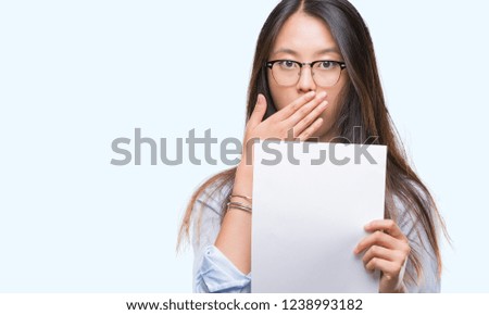 Young asian woman holding blank paper over isolated background cover mouth with hand shocked with shame for mistake, expression of fear, scared in silence, secret concept