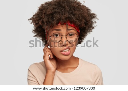 Puzzled uncertain black girl frowns face in displeasure, feels apathy, looks with uncertainty, cant find solution and way out of problem, has Afro haircut, models against white wall. Facial expression