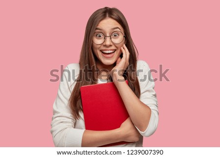 Positive European woman with straight hair, has toothy smile, giggles as has fun with classmates, wears round spectacles, holds red textbook, rejoices recieving good mark, models over pink wall