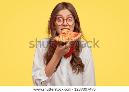 Emotive beautiful lady bites delicious pizza, looks directly at camera, has time for snack, visits pizzeria, surprised with low prices, models over yellow background. People, fast food and nutrition Royalty-Free Stock Photo #1239007495