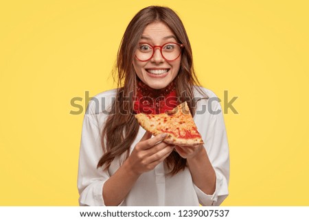 Photo of satisfied woman holds piece of pizza, feels pleased as spends free time with friends in pizzeria, looks happily directly at camera, wears casual outfit, isolated over yellow wall. Lunch Royalty-Free Stock Photo #1239007537
