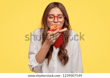 Mmm, so delicious! Dark haired pretty woman eats slice of Italian pizza, keeps eyes closed from pleasure, enjoys nice taste, wears glasses and shirt, isolated over yellow background. Eating concept Royalty-Free Stock Photo #1239007549