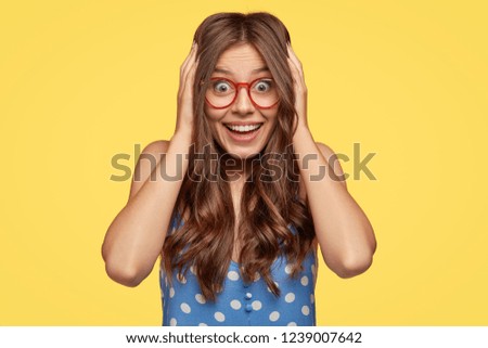 Excited happy lady listens awesome story, touches head with both hands, feels entertained, wears red rim spectacles and polka dot dress, stands against yellow background, expresses positive reaction