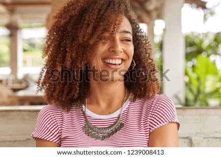 Headshot of funny black woman with curly hair, laughs at good joke, has toothy smile, shows white perfect teeth, wears striped casual t shirt, poses at terrace cafeteria. Afro American teenager indoor