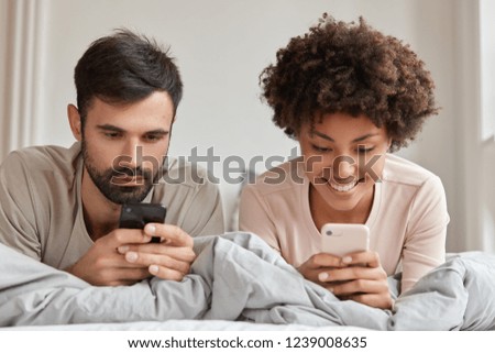 Photo of addicted woman and man of different races, use mobile phones in bed, connected to wireless internet, enjoy morning at weekend, have cheerful expressions. Modern technologies concept