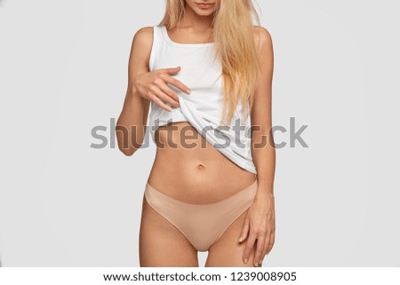 Unrecognizable blonde lady shows flat belly and slim waist, raises casual white vest, wears pants, demonstrates perfect slim body, stands against studio background. People, healthy lifestyle concept