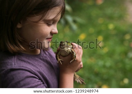funny child girl with frog in her hands, playing in nature and interest in wild animals. Summer day over garden