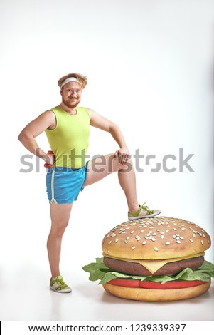 Funny picture of red haired, bearded, plump man on white background. Man wearing sportswear. Man put his leg to a huge sandwich