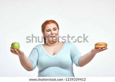 Funny picture of amusing, red haired, chubby woman on white background. Woman holding apple and sandwich. She is looking at a sandwich