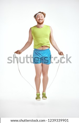 Funny picture of red haired, bearded, plump man on white background. Man wearing sportswear. Man holding skipping rope.