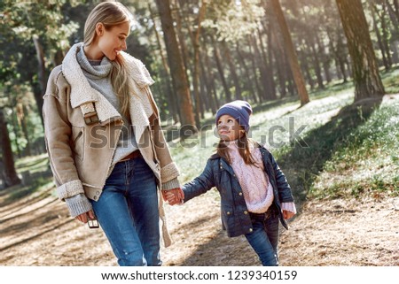 A little curly girl in hat is walking with her mother in woods. Cold season, bright sun is seen through the trees. Close-up
