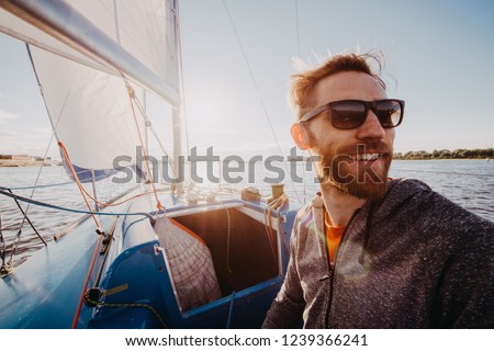 Man dressed in casual wear and sunglasses on a yacht. Happy adult bearded yachtsman close-up portrait. Handsome sailor on a boat smiling during regata on a sea or river. Royalty-Free Stock Photo #1239366241