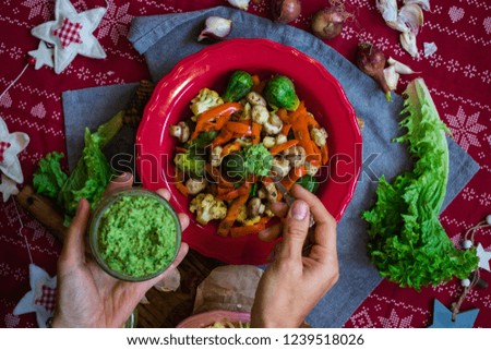 Woman hands adds spinach avocado dip sauce to baked roasted vegetables for Christmas lunch or dinner on red tablecloth. Vegan food with brussels sprouts, carrots, cauliflower, mushrooms. 