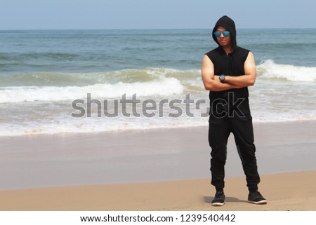 Handsome young man in sunglasses and sleeveless hoodie on beach during summer vacation