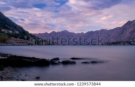 Long exposure sunset in bay of Kotor Montenegro.   Water appears flat with rocks in anchor, and puffy clouds, in a bay surrounded by jagged glowing mountain from sun setting on left side 